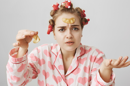 Indoor shot of clueless puzzled feminine girl in hair-curlers and pajamas, holding eye patch mask on forehead and in hand, having no clue how to apply it, gesturing over gray backgroundの写真素材