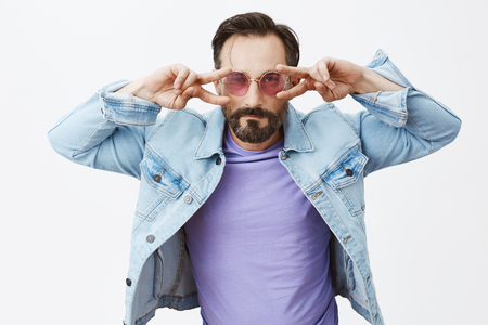 Drunk man going on dance floor, dancing from heart with passion and emotions, standing in denim jacket and purple sunglasses, showing peace or victory signs near eyes and gazing seriously at cameraの写真素材