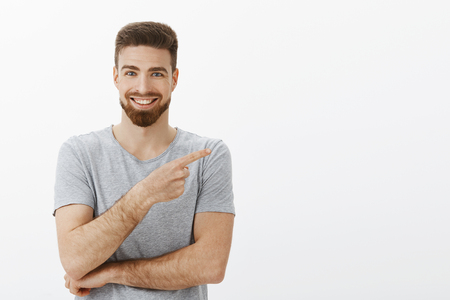 Good-looking charming independent entrepreneur with beard and cool hairstyle in grey t-shirt pointing right at copy space and smiling assured and delighted telling people about great productの写真素材