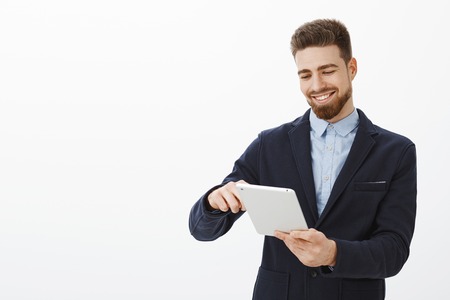 What pleasure looking at bank account full of money. Delighted handsome and successful businessman with beard and neat hairstyle in suit holding digital tablet smiling pleased looking at device screenの写真素材