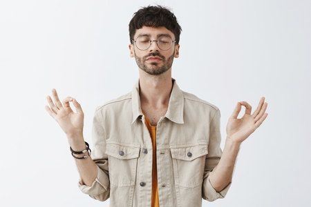 Calm good-looking and stylish urban guy with beard and trendy hairstyle in glasses meditating to relieve from stress being patient and relaxed practicing yoga with hands spread aside and closed eyesの写真素材