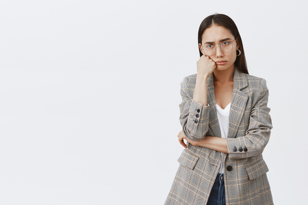 Portrait of moody sad girlfriend in fashionable outfit and glasses, leaning head on fist, sulking and frowning, expressing regret and disappointment, standing upset over gray backgroundの写真素材