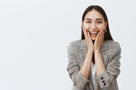 Studio shot of charming thrilled adult female entrepreneur, smiling broadly with intrigue, holding palms on cheeks, gazing with interest at camera while hearing amazing gossips, liking dirty rumorsの写真素材