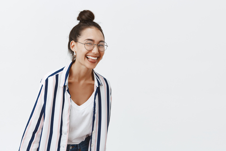 Girl just wanna have fun. Studio shot of charming playful woman leading active and happy lifestyle, bending towards camera while laughing over funny joke, standing in trendy outfit and glassesの写真素材