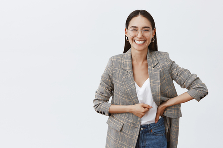 Stylish happy charming mongolian woman with bright optimistic smile, holding hand on waist and gazing with joy at camera, laughing over funny story, having fun during break in officeの写真素材