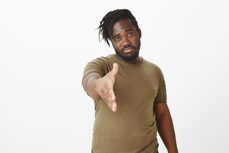 Indoor shot of confident serious-looking african guy with beard, pulling hand towards camera to give handshake, standing self-assured while greeting coworker or friend over greay backgroundの写真素材