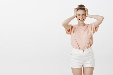 Woman suffering huge migraine, yelling from pain. Good-looking troubled girlfriend with freckles and bun hairstyle in stylish clothes, holding hands on head, screaming from painful feeling or regretの写真素材