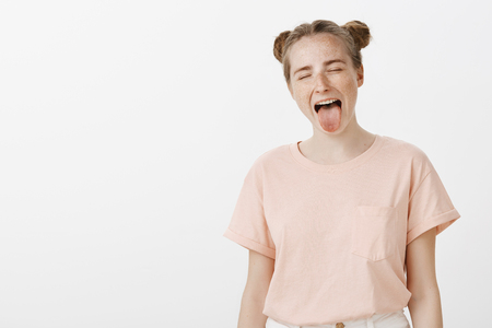 Horizontal shot of carefree childish girl having fun, being amused and entertained while sticking out tongue with closed eyes as if licking ice cream, standing in playful pose over gray backgroundの写真素材