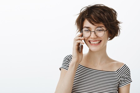 Happy you call. Portrait of happy positive female friend in round glasses and striped t-shirt, looking aside with joyful smile while holding smartphone near ear and talking with girlfriendの写真素材