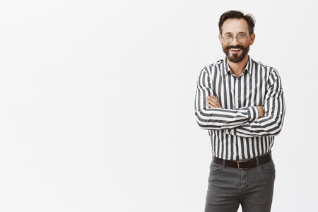 Attractive successful businessman in formal striped shirt and pants, holding hands crossed on chest and smiling broadly while standing on social evening, talking with business partners casuallyの写真素材