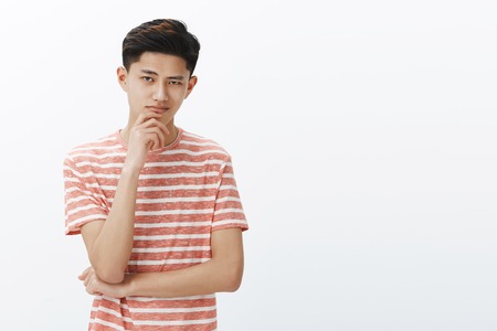 Guy knows what we need. Portrait of smart and creative good-looking determined young asian male student in striped t-shirt smirking self-assured standing in thoughtful pose with hand on chinの写真素材