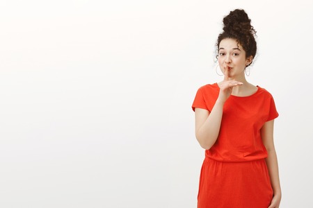 Portrait of creative cheerful Caucasian girlfriend with curly hair combed in bun, smiling curiously while saying shh, making shush gesture with index finger over mouth, making surprise over gray wallの写真素材