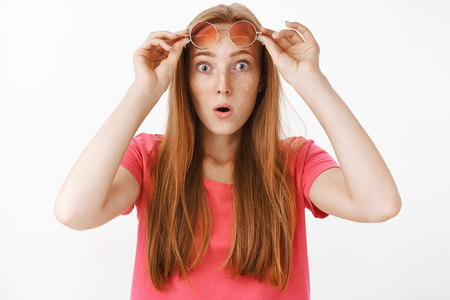 Waist-up shot of shocked and impressed speechless redhead female with freckles popping eyes and gasping with opened mouth taking off glassesの写真素材