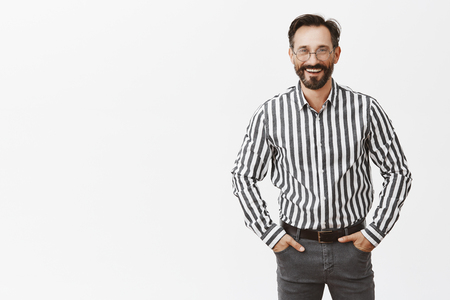 Portrait of good-looking friendly and happy kind man with beard in glasses holding hands in pockets and smiling broadly with confident lookの写真素材