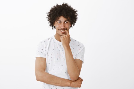 Man laughing over his own thought while thinking, smiling joyfully, holding finger on lip and gazing at upper right corner, imaging funny moment happened with boss, standing pleased over grey wallの写真素材