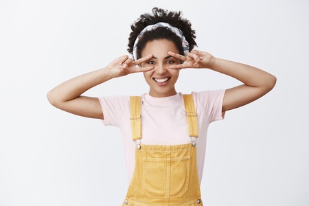 Peace friends. Attractive dark-skinned stylish female friend in headband over hair and yellow trendy overalls, showing peace gesture over eyes and smiling with carefree and happy expressionの写真素材