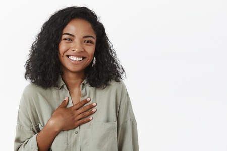 Waist-up shot of amused attractive and stylish young african american female with curly haircut being pleased receiving thanks being grateful for nice words holding palm on breast and smiling friendlyの写真素材
