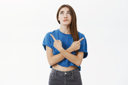Hmm girl having hesitations during making choice. Portrait of unsure gorgeous female in blue t-shirt biting lip looking up uncertain crossing hands against body poinding left and right over grey wallの写真素材
