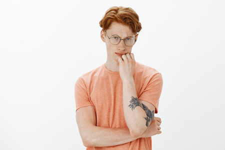 Indoor shot of serious handsome and thoughtful tattooed male with red hair and freckles in glasses, touching lip and frowning while being focused on math quiz, standing and thinking over gray wallの写真素材