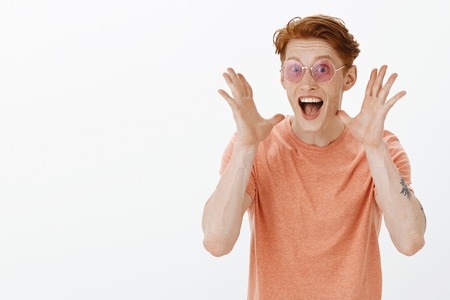 Good-looking stylish party man with ginger hair and freckles, smiling from excitement and joy while explaining incredibly great news, gesturing and shaking palms, standing in sunglasses over grey wallの写真素材