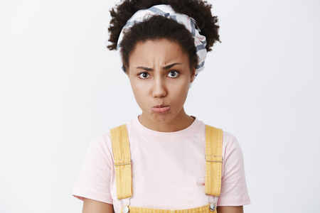 You promised to spend time with me. Portrait of upset and offended sad african american female in headband and yellow dungarees, pursing lips, frowning and sulking from offence over grey wallの写真素材