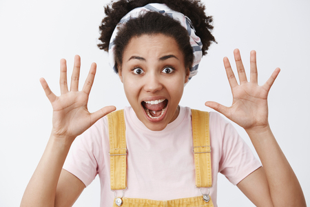 Boo. Cute happy elder sister with dark skin in trendy yellow dungarees and headband raising palms and shouting, wanting scare sibling, crowling from back, standing childish over gray backgroundの写真素材
