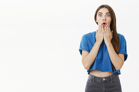Indoor shot of amused shocked and speechless cute european brunette female in blue t-shirt gasping from excitement holding palms on chin amused and surprised staring at camera amazed against grey wallの写真素材