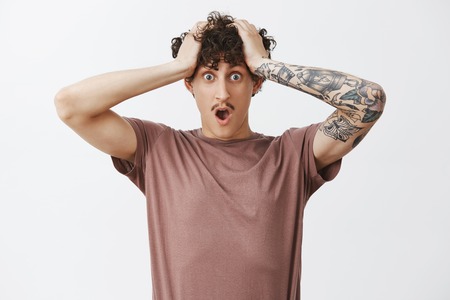 Time playing against guy. Shocked and worried concerned good-looking young male with moustache and curly hairstyle holding hands on head dropping jaw being in trouble and perplexed situationの写真素材