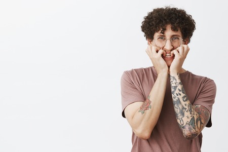 Studio shot of intense scared and timid good-looking funny european guy with moustache curly hair and tattoos biting fingrnails and frowning feeling frightened of scary movie on tvの写真素材