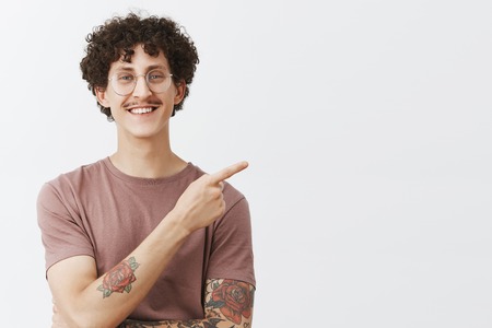 You would not find better place for advertising. Friendly handsome and creative young stylish male model with curly hairstyle tattoos and moustache smiling joyfully pointing at upper right cornerの写真素材
