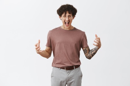 Guy getting pissed off on work. Fed up stylish creative male designer in fury yelling clenching raised fists in anger staring with hate and outrage at camera standing irritated over gray backgroundの写真素材