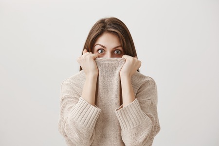 Woman hears terrifying story making her tremble from fear. Portrait of cute adult caucasian girl hiding in sweater, peeking out of collar with popped eyes being scared or shocked over gray wallの写真素材