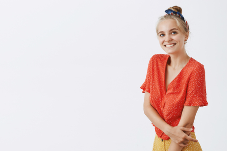 Studio shot of charming emotive and cute timid and shy girl with blond hair combed with headband wearing trendy polka-dot blouse, smiling and touching armの写真素材