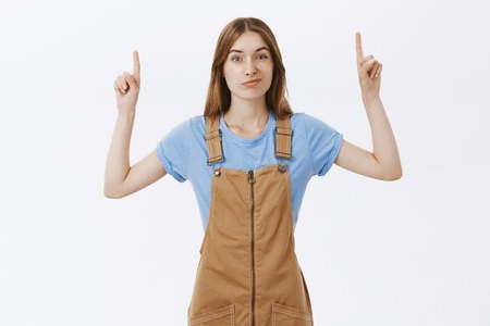 Doubtful and hesitant cute unimpressed girlfriend in brown overalls over t-shirt raising hands pointing up and smirking with displeased suspicious expression giving negative feedback over white wallの写真素材