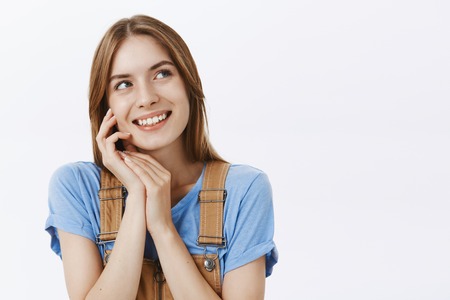 Dreamy cute and feminine charming european female in blue t-shirt and overalls touching face gently with palm smiling delighted and nostalgic looking joyfully at upper right corner, dreamingの写真素材
