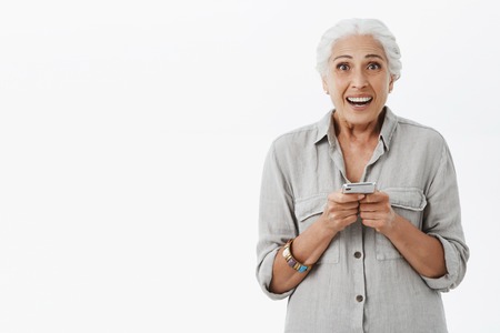 Portrait of excited and pleased charming granny with grey hair in shirt holding telephone gazing...
