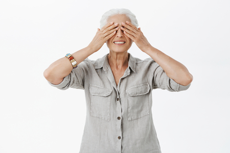 Portrait of charming hopeful and charismatic senior woman playing with kids in hide and seek closing eyes with palms and smiling joyfully against gray backgroundの写真素材