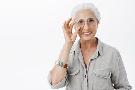 Waist-up shot of charming kind and nice granny in sight glasses touching rim of eyewear and smiling...