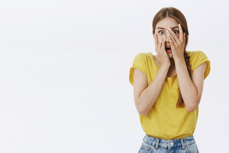 Waist-up shot of surprised young and cute slender woman with braid opening mouth from amazement closing face with palms peeking through fingers with anticipation and astonishment over white wallの写真素材