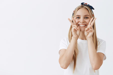 Waist-up shot of charming friendly-looking young blond woman in headband showing victory or peace signs on face and smiling joyfully, posing for photograph in cute way over gray backgroundの写真素材