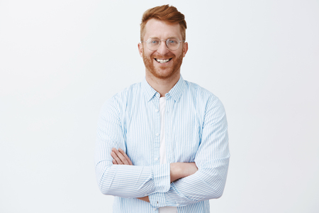 Redhead guy knows everything about business, standing in trendy shirt over gray wall with crossed hands on chest, smiling joyfully and gazing at camera while taking part in conversation with partnersの写真素材