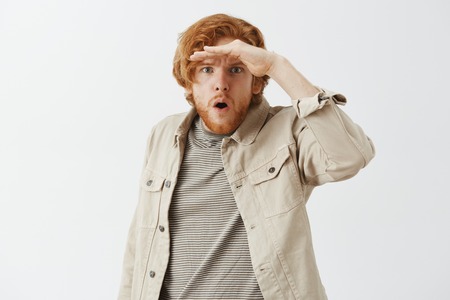 Studio shot of amazed and stunned male redhead with beard holding palm on forehead as if looking on distance opening mouth while looking far awayの写真素材