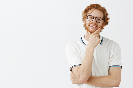Thoughtful handsome young genious in glasses with red hair standing in thinker pose with hand on chin gazing with smart and curious expression, observing interesting and intriguing conceptの写真素材
