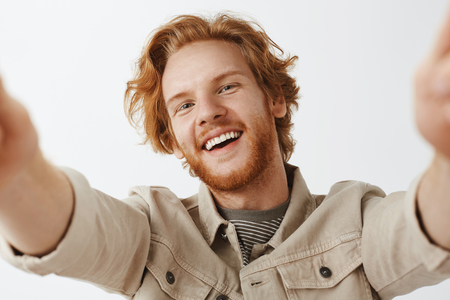 Close-up shot of charming friendly and carefree happy redhead guy with stylish wavy hairstyle and beard tilting from joy smiling, pulling hands towards camera to take selfieの写真素材