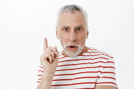 Excited creative and smart wise senior man with grey hair and beard in striped t-shirt raising index finger in eureka gesture folding lips, talking, adding suggestion over white backgroundの写真素材