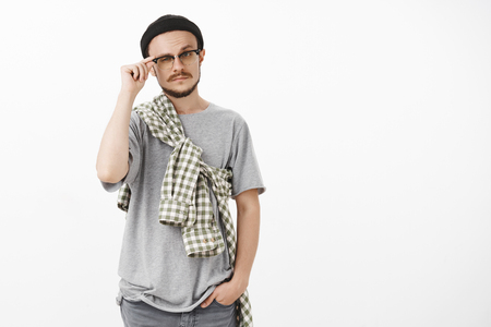 Studio shot of smart artistic handsome male with beard and moustache raising eyebrow and squinting touching rim of glasses standing curious and intrigued in beanie and glasses over white backgroundの写真素材