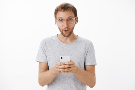 Portrait of impressed handsome  guy with bristle saying wow with pleased grin holding smartphone wearing earbudsの写真素材