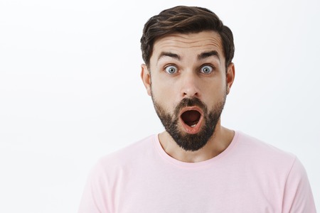 Close-up shot of impressed and shocked stunned caucasian guy with beard dropping jaw from amazement popping eyes at camera being surprised and excited with shocking news over gray backgroundの写真素材