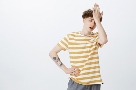 Oh gosh why me. Portrait of uneasy bothered and fed up young teenage boy with curly hairstyle and tattoo on arm punching forehead with palm looking drained and tired as standing over gray wallの写真素材