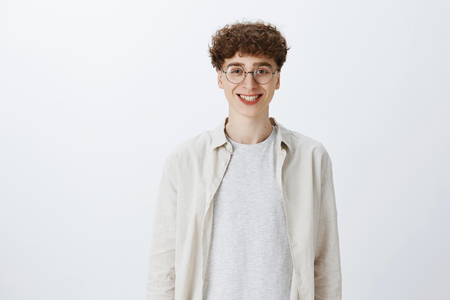 Waist-up shot of optimistic and happy young handsome boy in round glasses with curly hairstyle smiling broadly as standing over gray background carefree and enthusiasticの写真素材